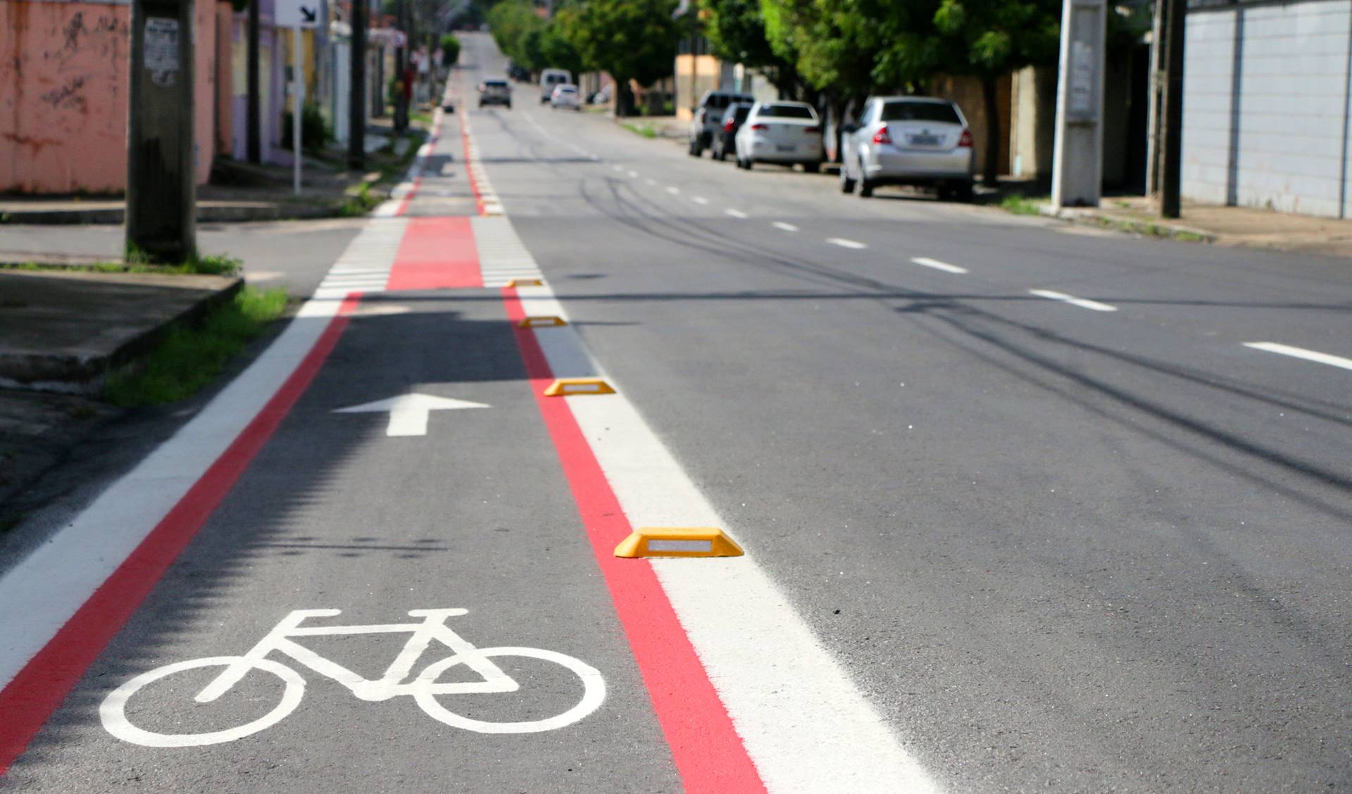 a imagem mostra a sinalização de uma ciclofaixa, que consiste em linhas retas brancas e vermelhas, demarcando o espaço na lateral da rua e do desenho branco de uma bicicleta